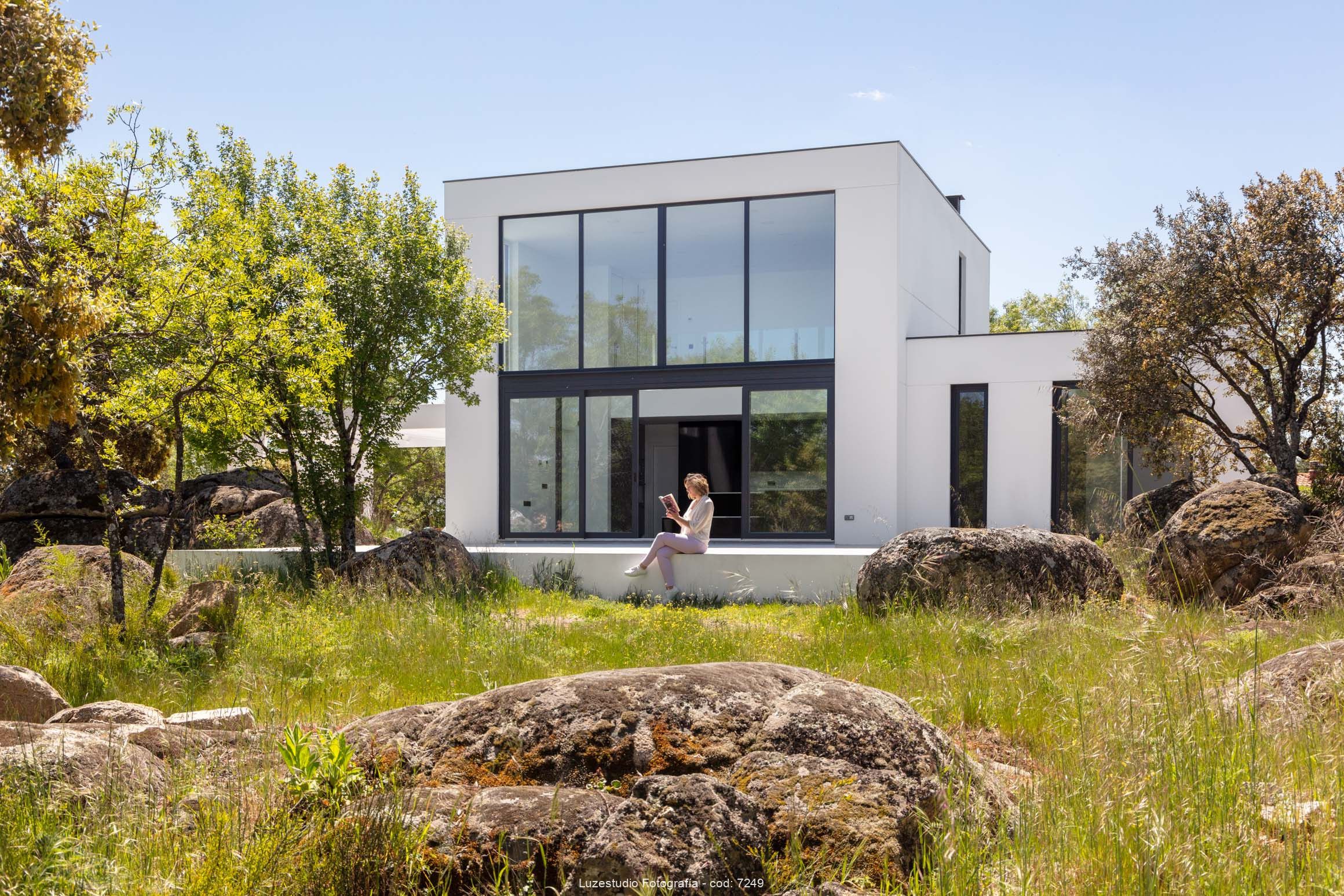 foto de arquitectura con mujer joven leyendo un libro sentada en el porche de una casa modular de hormigón rodeada de naturaleza salvaje con piedras y encinas